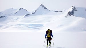 A person wearing winter gear and carrying a backpack skis across a vast, snowy Antarctic landscape toward jagged, snow-covered mountains under a pale sky, embodying the spirit of Antarctic Logistics and Expeditions.