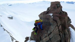 A climber with a large backpack ascends a rugged, snow-dusted rock formation surrounded by vast, snow-covered mountains and valleys on an Antarctic Logistics and Expeditions journey.