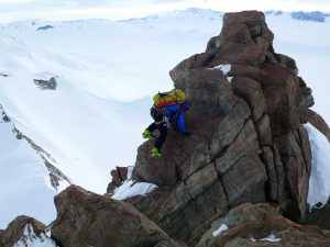 Two climbers with helmets and gear ascend a steep, rocky peak surrounded by snow-covered mountains under a cloudy sky. Connected by rope, they navigate the rugged terrain—a testament to Antarctic Logistics and Expeditions in remote Antarctica.