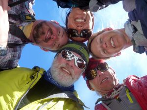 Five people in outdoor clothing stand in a circle, smiling down at the camera with blue Antarctic sky behind them. Some wear sunglasses and hats, suggesting an Antarctic Logistics and Expeditions adventure near the South Pole.