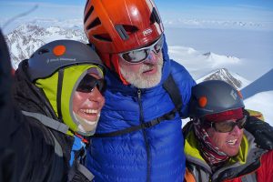 Three climbers wearing helmets, sunglasses, and winter gear smile and embrace at a snowy mountain summit in Antarctica, with a vast, icy landscape and distant peaks visible behind them under a clear sky.