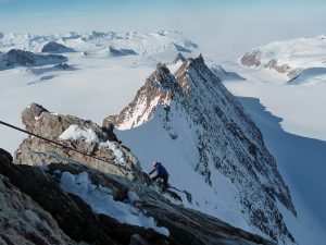 Two climbers ascend a snowy, jagged mountain ridge in Antarctica, surrounded by vast glaciers and snow-covered peaks under a clear sky. The landscape stretches into the distance, capturing the remote, icy wilderness near the South Pole.