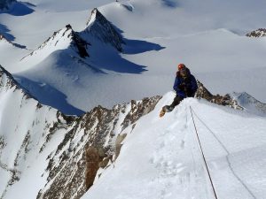 A climber in winter gear ascends a narrow, snowy mountain ridge with ropes, surrounded by steep snow-covered peaks and deep valleys under a clear sky—capturing the spirit of Antarctica and Antarctic Logistics and Expeditions.