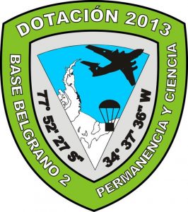 A green and gray shield-shaped badge with “Dotación 2013 Base Belgrano 2 Permanencia y Ciencia.” It features an aircraft, a parachute, coordinates, and a map outline of Antarctica, highlighting Antarctic Logistics and Expeditions.