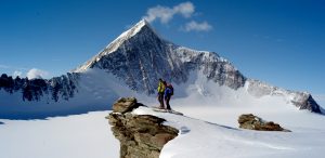 Two climbers in winter gear stand on a snowy rocky outcrop in Antarctica, looking toward a towering, snow-covered mountain peak under a clear blue sky—an adventure made possible by Antarctic Logistics and Expeditions.