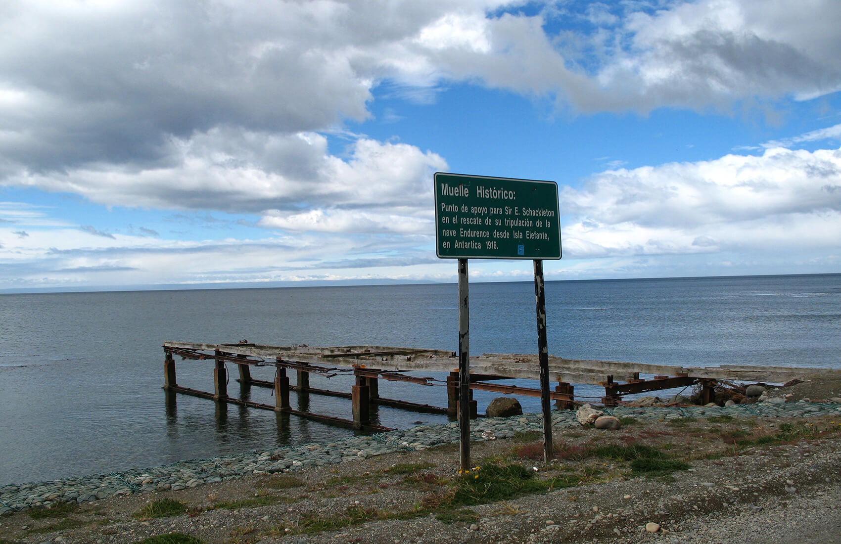 A weathered wooden pier extends into a calm sea under a cloudy sky. In the foreground, a green historical sign in Spanish stands beside the pier on a rocky shoreline, recalling early Antarctic Logistics and Expeditions to the South Pole.