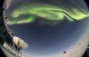A vibrant green aurora sweeps across a starry night sky above an Antarctic research station, with snow-covered ground and red flags marking the landscape in Antarctica near the South Pole.