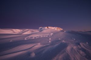 A snow-covered landscape at dusk with soft pink and purple lighting. In the distance, a flat structure partially buried by snow sits under a clear, darkening sky, evoking the remote beauty of Antarctica near the South Pole.