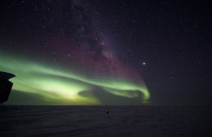 A night sky filled with stars and the Milky Way, vibrant green aurora borealis lights above a snowy, dark Antarctic landscape. A building from Antarctic Logistics and Expeditions is partially visible on the left side of the image.