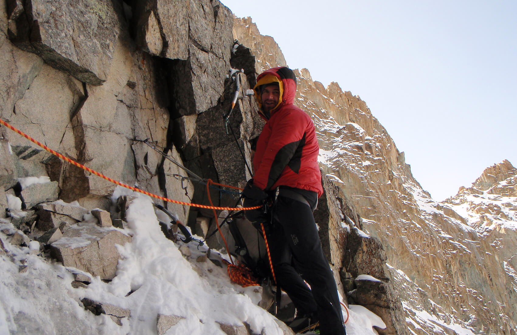 A climber in a red jacket and helmet is secured with ropes on a snowy, rocky mountain slope in Antarctica, with steep cliffs and a clear sky in the background.