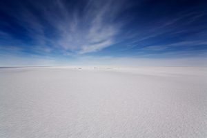 Vast, flat expanse of white salt flats under a dramatic blue sky with wispy clouds in Antarctica, with a faint line of distant dark objects on the horizon.