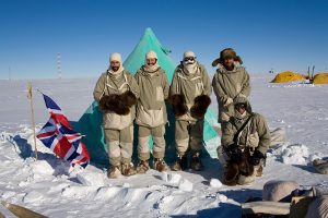Five people in thick, fur-trimmed Arctic clothing stand in front of a turquoise tent on snowy ground near an Antarctic Logistics and Expeditions camp, with a British flag and yellow tents behind them under the clear blue sky of Antarctica.