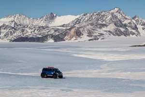 A blue 6-wheel vehicle drives across a vast, icy glacier in Antarctica, with rugged, snow-covered mountains in the background under a clear blue sky.