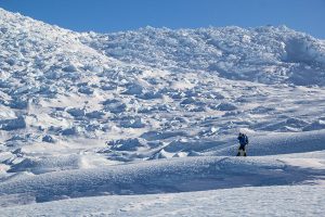 A lone mountaineer wearing winter gear stands on a snow-covered slope in Antarctica, surrounded by vast, rugged, icy terrain with a backdrop of large snowdrifts under a clear blue sky—an adventure supported by Antarctic Logistics and Expeditions.