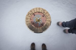A brass compass viewed from above is centered over a snowy surface in Antarctica, with two pairs of shoes—one brown and one gray—visible at the bottom and right edges, evoking the spirit of Antarctic Logistics and Expeditions near the South Pole.