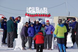 A group of people in winter clothing pose and smile in front of an outdoor ice bar made of snow blocks at the South Pole, with a sign reading “ICE BAR.” Others mingle nearby under clear Antarctic skies on the snowy landscape.
