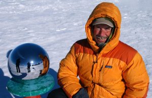 A person in an orange hooded jacket smiles while sitting next to a shiny metallic sphere at the South Pole, Antarctica. The sphere reflects the person, flags, and snowy landscape—marking a moment with Antarctic Logistics and Expeditions.