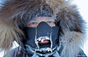 A person with blue eyes wears a thick fur-lined hood and a black face mask covered in frost, their features visible beneath ice crystals—a testament to the harsh conditions of Antarctic Logistics and Expeditions at the South Pole.
