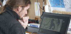 A person with long hair in a ponytail looks thoughtfully at a laptop displaying a detailed map of Antarctica. Maps and charts, including those related to Antarctic Logistics and Expeditions, are posted on the wall behind them in a workspace.