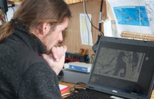 A person with long hair and a beard looks thoughtfully at a map of Antarctica displayed on a Dell laptop, surrounded by papers and notes about Antarctic Logistics and Expeditions pinned on the wall in a workspace.