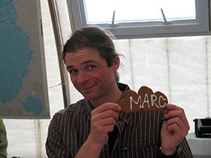 A person with long hair in a striped shirt smiles and holds a brown cookie with MARC written in white icing. Indoors in Antarctica, they stand before a map and window, capturing the spirit of Antarctic Logistics and Expeditions.
