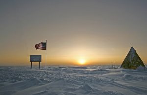 A snowy landscape at sunset near the South Pole with an American flag, a sign, and a tent on the right. The sun is low on the horizon, casting golden light over Antarctica’s snow-covered ground—likely part of Antarctic Logistics and Expeditions.