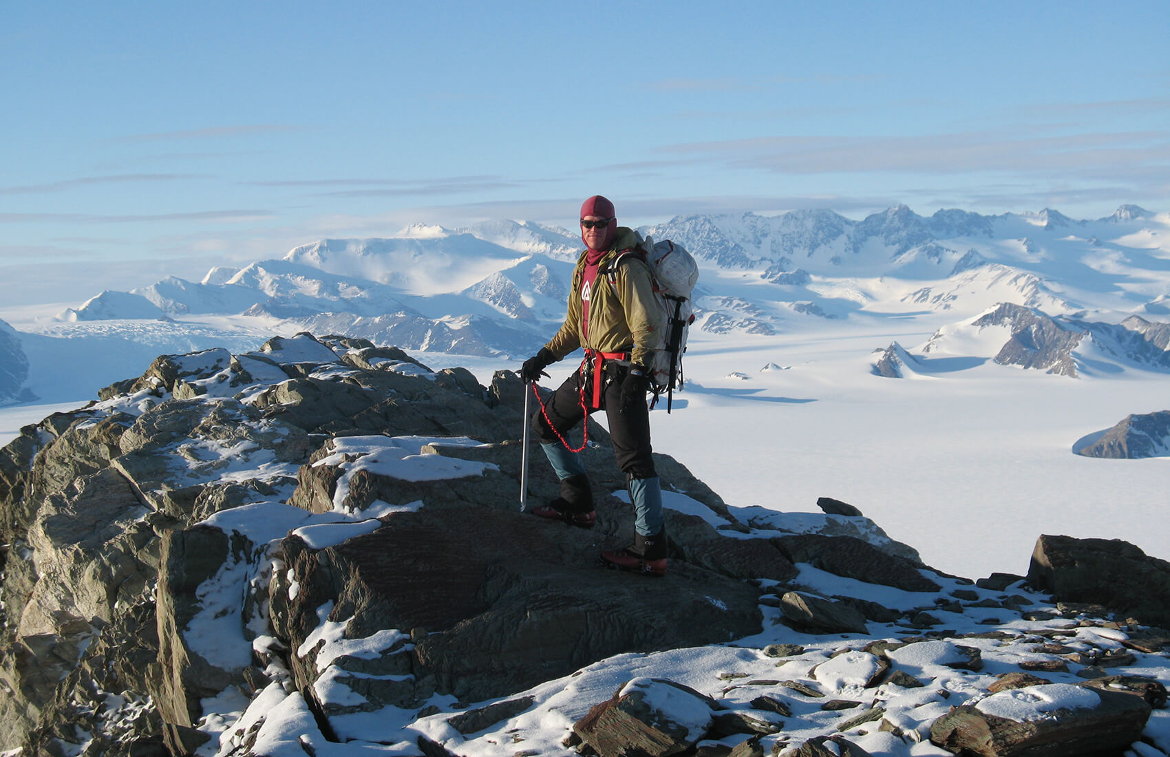 A climber in winter gear stands on a rocky, snow-covered Antarctic peak, holding an ice axe. Snowy mountains and an expansive glacier stretch toward the horizon under a clear blue sky, capturing the spirit of Antarctic Logistics and Expeditions.