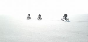 Three cyclists with backpacks ride single file across a vast, snowy Antarctic landscape under bright, overexposed light—a scene reminiscent of expeditions supported by Antarctic Logistics and Expeditions, their faint shadows stretching behind them.