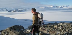 A person wearing outdoor gear and carrying a backpack stands on rocky terrain, gazing over Antarctica’s vast, snowy landscape with mountains in the distance under a partly cloudy sky—an adventure supported by Antarctic Logistics and Expeditions.