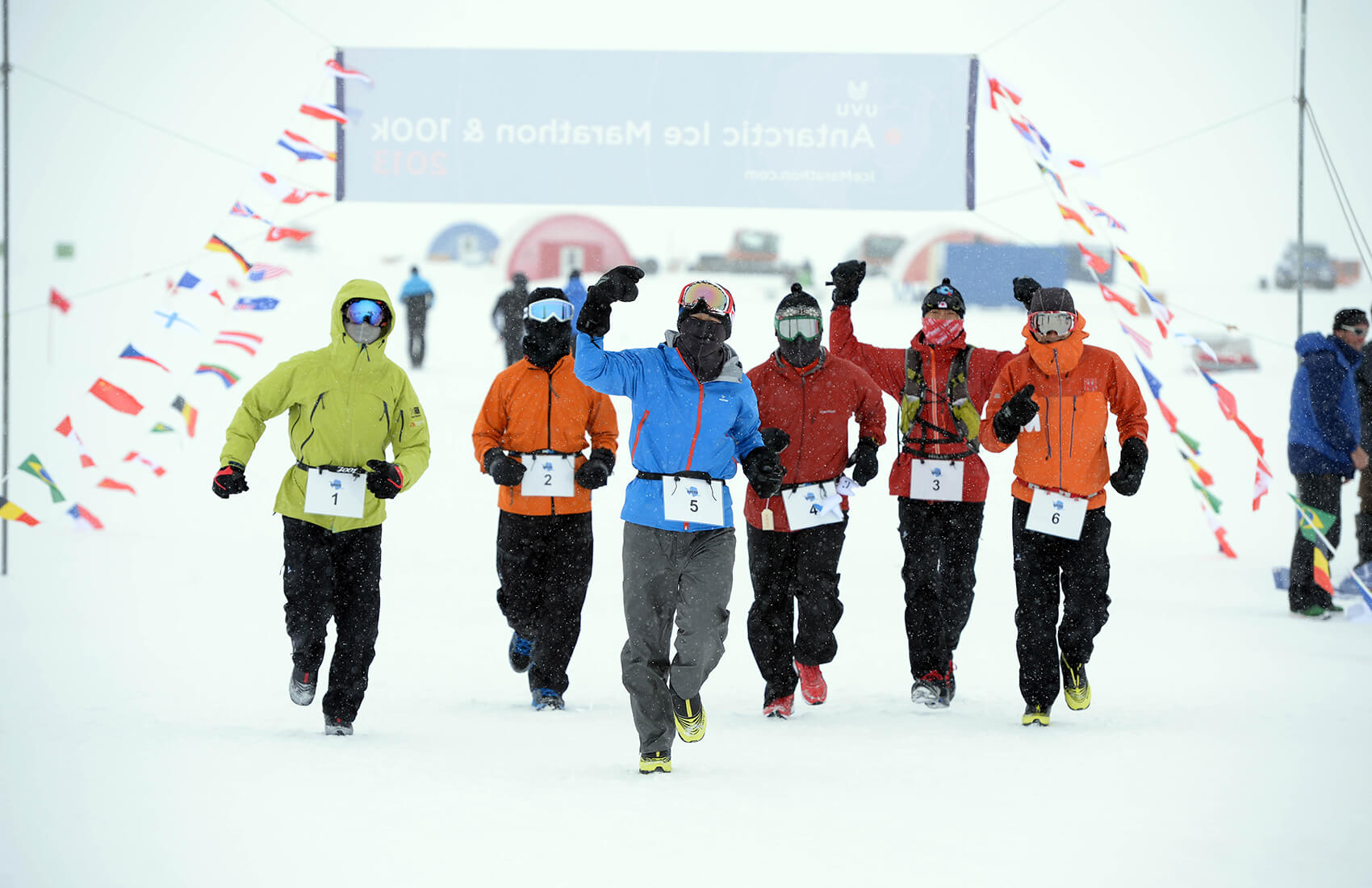 A group of six runners in winter gear and numbered bibs race toward the finish line at an Antarctic Logistics and Expeditions marathon event, with flags and tents dotting the snowy expanse of Antarctica in the background.