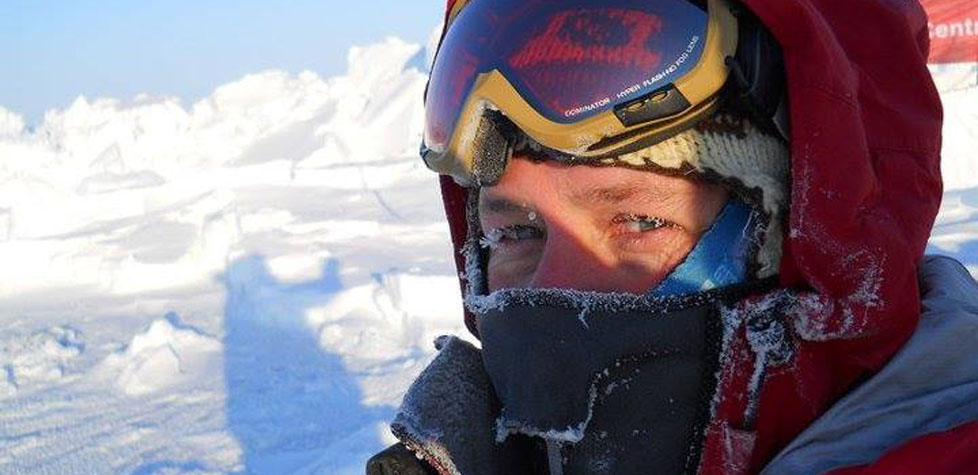 Bundled in winter gear with a frost-covered face, ski goggles, hat, and scarf, a person stands in a sunlit Antarctic landscape with icy formations—epitomizing the spirit of Antarctic Logistics and Expeditions.