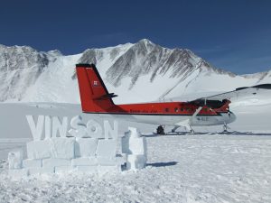 A red and white airplane is parked on snow in Antarctica, with snowy mountains in the background. In the foreground, VINSON is spelled out in large letters made of blocks of ice or snow.
