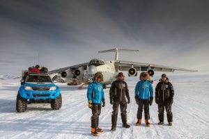 Four people in winter gear stand on snow beside a blue off-road vehicle and a large cargo plane, with Antarctic Logistics and Expeditions at the South Pole, framed by mountains and a cloudy sky in Antarctica.