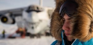 A person in a blue parka with a fur-lined hood and goggles stands in the foreground, while an Antarctic Logistics and Expeditions plane is parked on a snowy surface in the blurred background of Antarctica.
