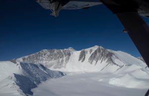 View from an aircraft over a vast, snow-covered mountain range in Antarctica with rugged peaks and deep valleys under a clear blue sky. Part of the Antarctic Logistics and Expeditions aircraft's wing and landing gear are visible above.