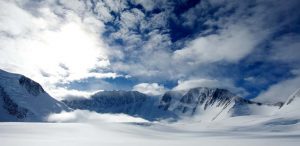 Snow-covered mountains in Antarctica rise beneath a dramatic, partly cloudy sky as sunlight filters through, illuminating the vast, white landscape—a breathtaking scene reminiscent of journeys with Antarctic Logistics and Expeditions.