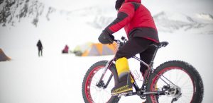 A person in a red jacket and yellow boots rides a fat-tire bike across an Antarctic snowy landscape with tents and mountains in the background, supported by Antarctic Logistics and Expeditions. Other people are visible in the distance.