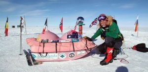 A person in cold-weather gear kneels on snow beside a sled shaped like pink breasts, decorated with flags and sponsor logos from Antarctic Logistics and Expeditions. Several international flags stand in the background of sunny Antarctica.