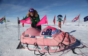 A person in cold-weather gear stands next to a pink sled covered in flags and sponsor logos on a snowy Antarctic landscape, with various country flags, a reflective globe marker, and the South Pole nearby.