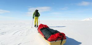 A person in bright winter gear pulls a sled loaded with red bags across the vast, snowy landscape of Antarctica under a clear blue sky, showcasing the adventurous spirit of Antarctic Logistics and Expeditions.