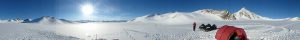 Panoramic view of a snowy mountain landscape under a clear blue sky in Antarctica, with sun shining brightly, snowmobiles parked on the right, and a person in red near a tent—an adventure supported by Antarctic Logistics and Expeditions.