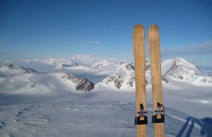 Two wooden skis stand upright in the snow on a mountain, overlooking a panoramic view of snow-covered peaks and a clear blue sky reminiscent of Antarctica's vast beauty—perfect for an Antarctic Logistics and Expeditions adventure.