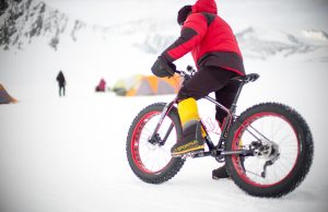A person in a red jacket rides a fat-tire bike on snowy terrain near several tents, with Antarctica’s snow-covered mountains in the background and another person walking nearby—capturing the spirit of Antarctic Logistics and Expeditions.