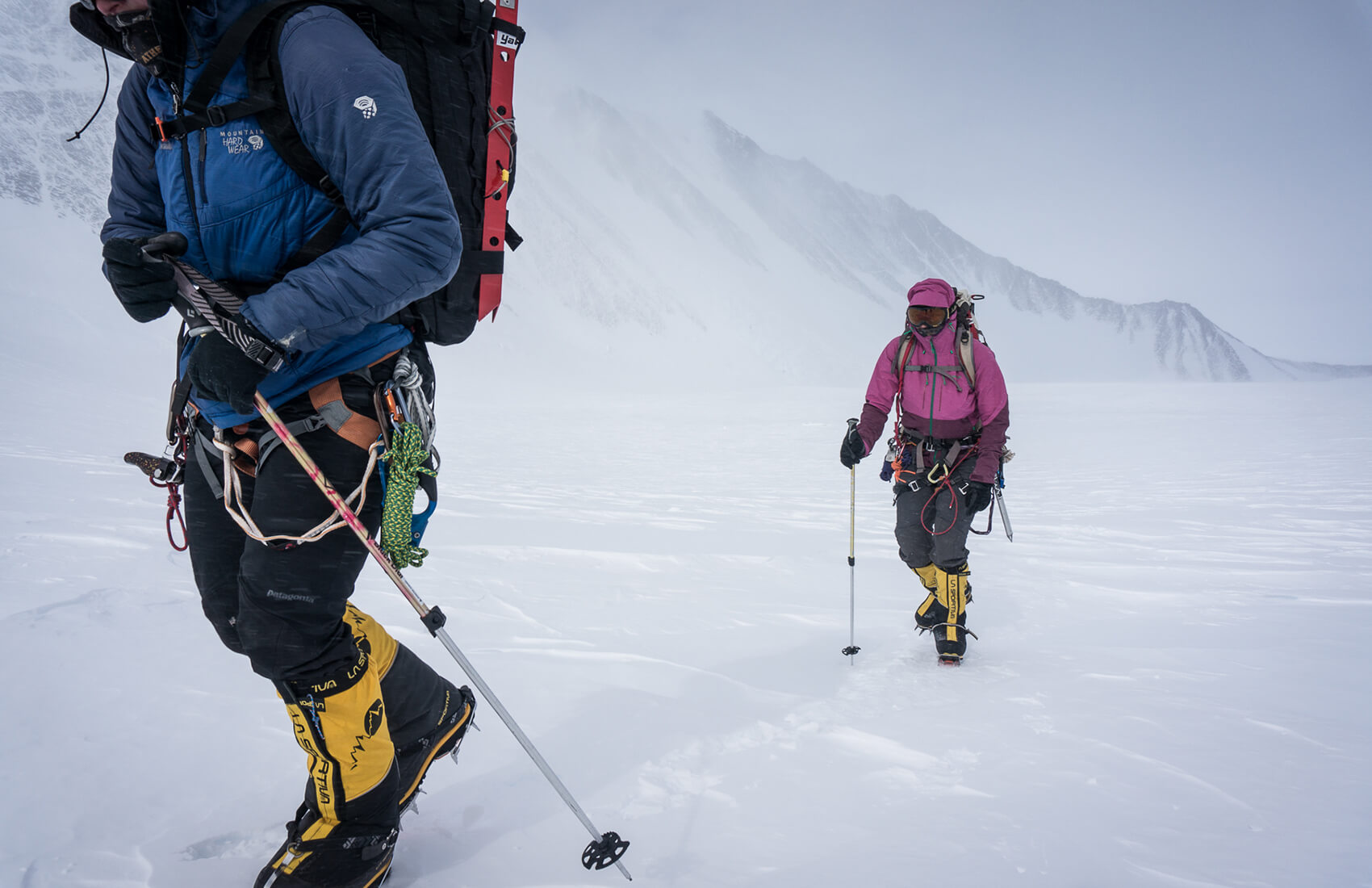 Two climbers in heavy winter gear and snow boots trek through a snowy, windswept mountain landscape in Antarctica, using trekking poles for support. The background shows rugged, snow-covered peaks under a cloudy sky—typical of an Antarctic Logistics and Expeditions journey.