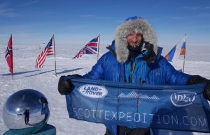 A person in a blue parka stands on snow at the South Pole beside a reflective sphere, smiling and holding a “ScottExpedition.com” banner. Antarctic Logistics and Expeditions flags from Norway, the USA, the UK, and Russia fly in the clear sky background.