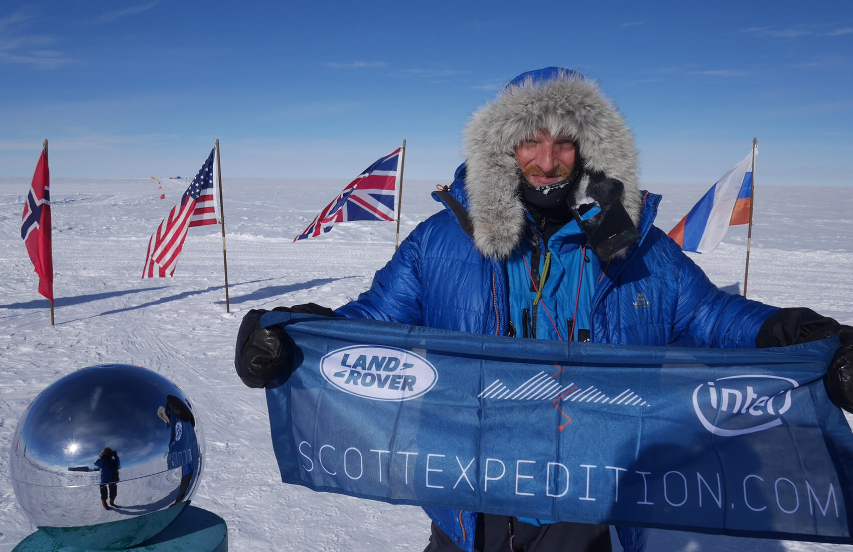 A person in a blue parka stands on snow at the South Pole beside a reflective sphere, smiling and holding a “ScottExpedition.com” banner. Antarctic Logistics and Expeditions flags from Norway, the USA, the UK, and Russia fly in the clear sky background.
