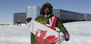 A person in a green insulated jacket and fur-lined hood stands on snow in Antarctica, smiling and holding a Welsh flag, with a modern, grey research station—supported by Antarctic Logistics and Expeditions—in the background under a clear blue sky.