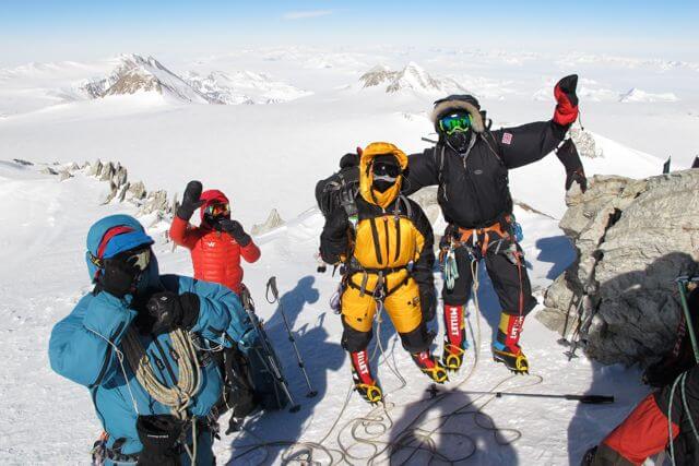 Four climbers dressed in colorful, heavy winter gear stand on a snowy mountain summit in Antarctica, surrounded by ropes and equipment. Snow-covered peaks stretch behind them as two climbers pose with arms raised, celebrating their Antarctic Logistics and Expeditions adventure.