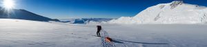 A lone skier with a backpack and sled journeys across a vast, snowy landscape under a clear blue sky, sunlight gleaming on distant snow-covered mountains—an adventure reminiscent of Antarctica and South Pole expeditions.