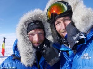 Two people in blue winter parkas with fur-lined hoods take a close-up selfie in the snowy landscape of Antarctica. A blurry flag stands in the background beneath a clear South Pole sky, hinting at their Antarctic Logistics and Expeditions journey.